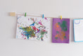 Load image into Gallery viewer, A wall-mounted art display featuring kids art work with colorful mini clothes pegs and natural jute twine hanging line.