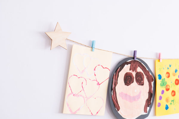 A wooden star art display mounted on a wall with clothes pegs holding up pieces of children's artwork.