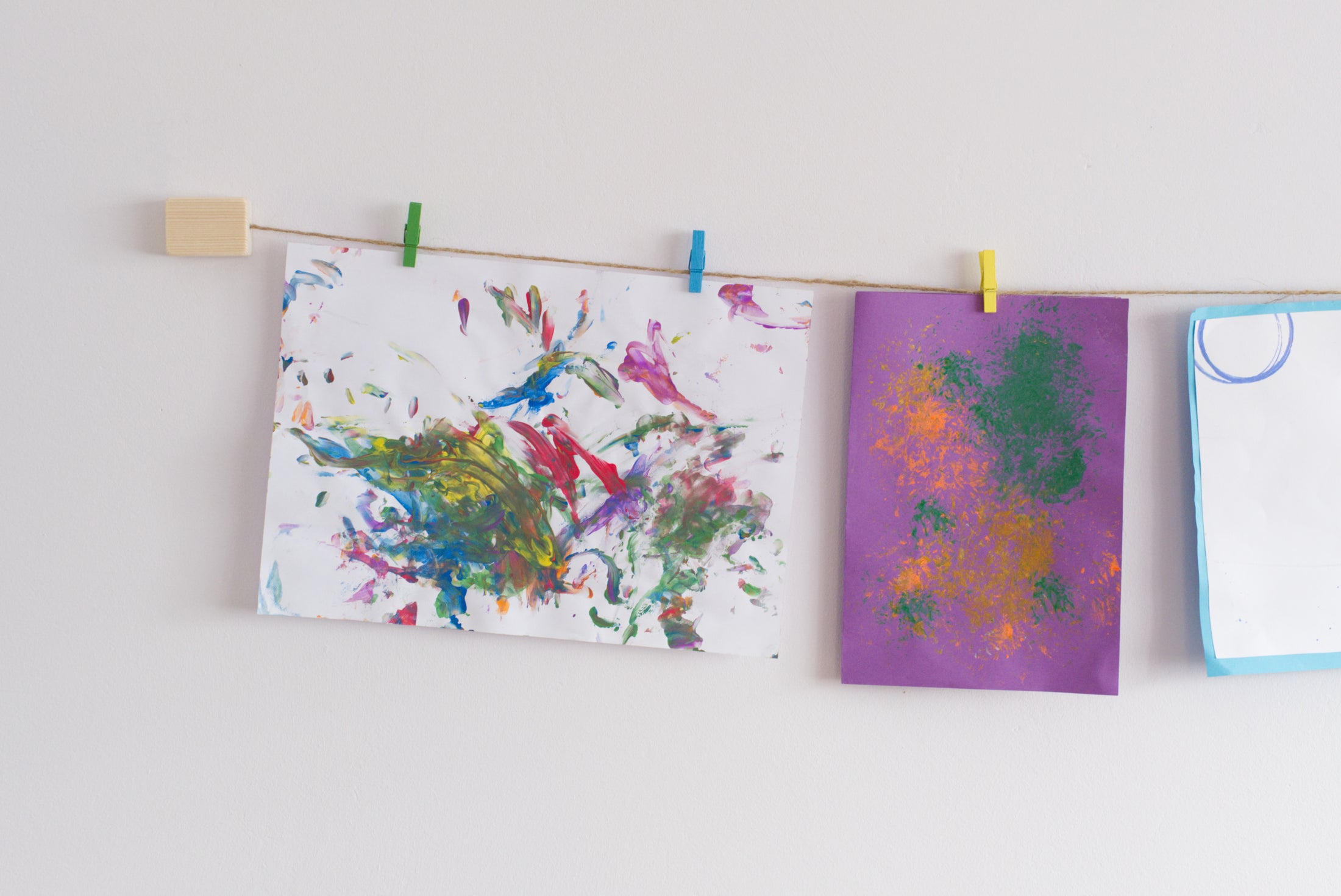 A wall-mounted art display featuring kids  art work with colorful mini clothes pegs and natural jute twine hanging line.